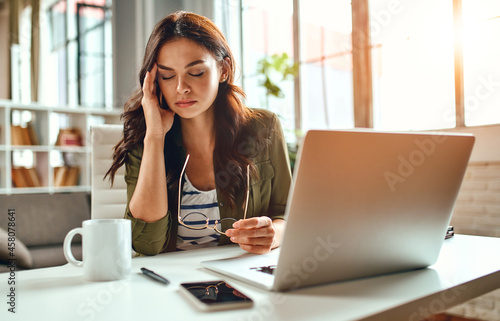 Tired business woman in stress works at a laptop while sitting at a table at home and holds her hand on her temples, migraine attack. Freelance, work from home.