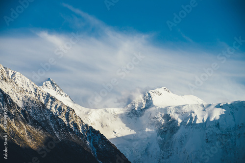 Atmospheric alpine landscape with high snowy mountain with peaked top under cirrus clouds in sky. Low cloud on big snow covered mountain in sunshine. Black rocks and white-snow pointy peak in sunlight
