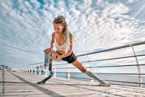 Image of disabled athletic girl in sportswear doing sit ups and stretching bionic leg on the bridge near sea.