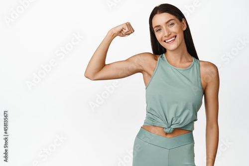 Smiling brunette fitness girl showing muscles on arm, flexing biceps and looking happy at camera, workout in gym, wearing sportswear, white background