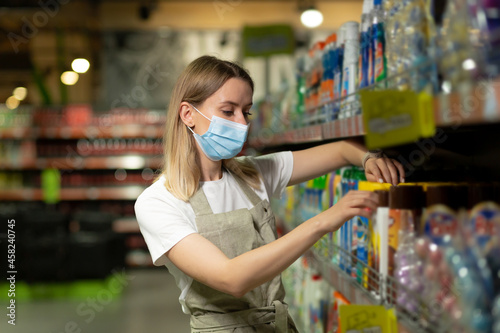 portrait of saleswoman, in protective face mask woman smiling and looking at camera in supermarket. Pleasant friendly female seller standing in the store between the rows. worker with crossed arms