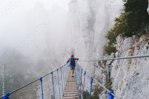 A young man in mountaineering equipment walks uphill on a suspension bridge in the fog. Suspension bridge on Mount Ai-Petri, Yalta, Crimea. The concept of travel, active lifestyle, tourism.