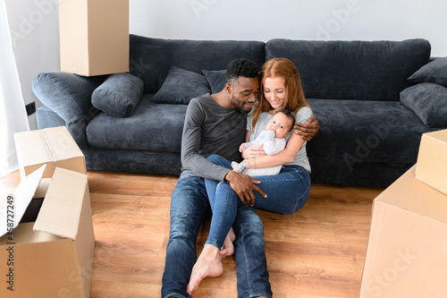 Happy mixed-race international family during unpacking day, moving in the new home. African father, caucasian mother and cute biracial baby daughter lying on the floor among cardboard boxes