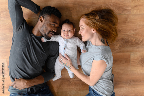 Happy mixed-race international family of three lying on the floor, happy black husband, caucasian wife and their biracial baby girl looking at the camera, top view