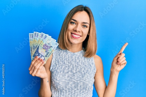 Young caucasian blonde woman holding 100 romanian leu banknotes smiling happy pointing with hand and finger to the side