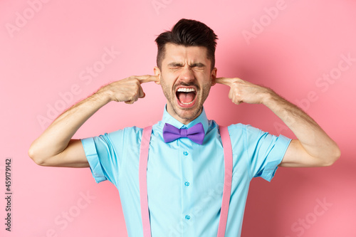 Irritated young man block noise, shut ears with hands and screaming annoyed, being disturbed by loud sound, standing over pink background