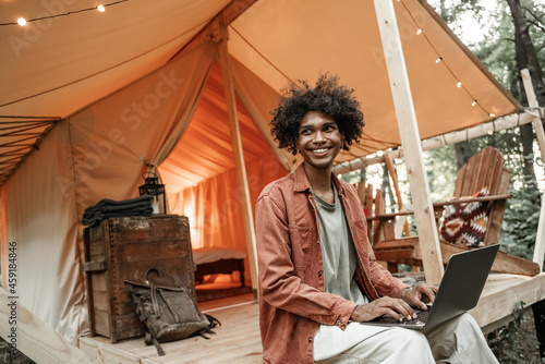 Young smiling African man with piercing sitting at glamping typing on laptop. Camping lifestyle. Low budget travel, holiday. Wi-fi connection information communication technology. Remote work