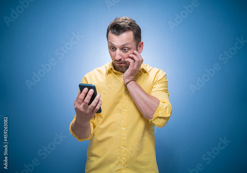 Astonished guy staring to phone over blue background, dresses in yellow shirt. Trendy man. Unbelievable message
