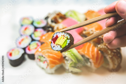 Man takes Tasty Colorful assorted Set of different type Sushi with chopsticks. Dinner in Japanese style. Healthy food. Filadelfia and Maki sushi rolls with Avocado, Tuna, Salmon, fish and Prawns.