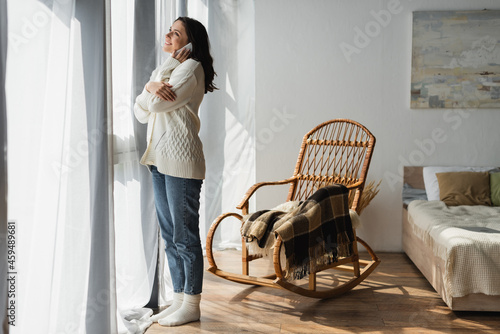 full length view of woman in jeans and white cardigan calling on smartphone near window and wicker chair
