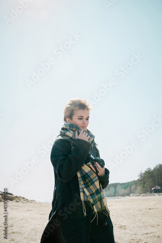 Young woman in a coat and scarft with camera standing on a beach in cold weather, selective focus