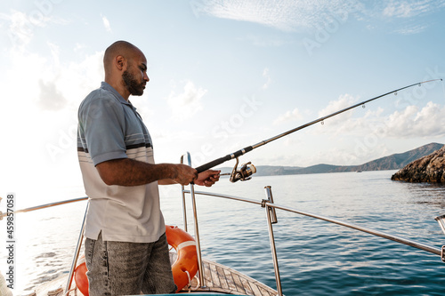 Young african american man standing with fishing rod on a sailboat fishing in open sea on sunset