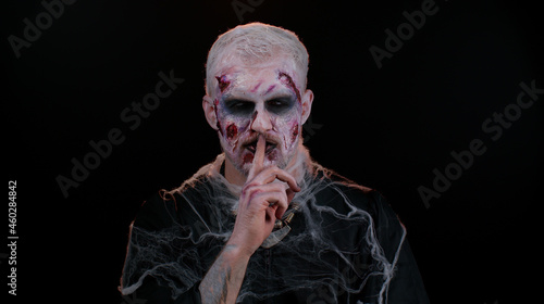 Shh be quiet. Sinister man with horrible scary Halloween zombie makeup in costume presses index finger to lips makes silence gesture sign do not tells secret. Dead guy with wounded bloody scars face