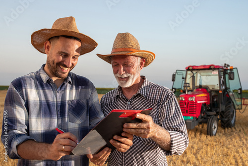 Young farmer and senior agronomist signing documents in field with machinery in background.