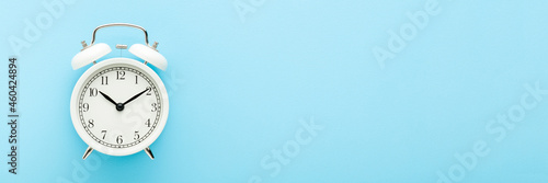 White alarm clock on light blue table background. Pastel color. Closeup. Wide banner. Time concept. Empty place for text. Top down view.