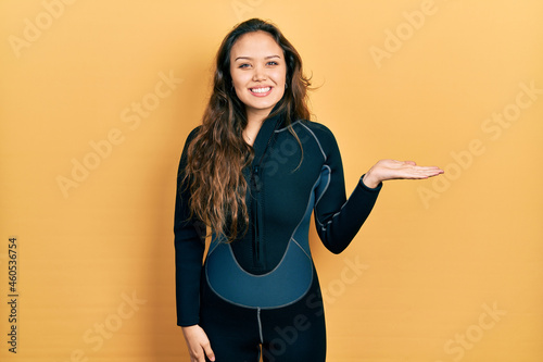 Young hispanic girl wearing diver neoprene uniform smiling cheerful presenting and pointing with palm of hand looking at the camera.