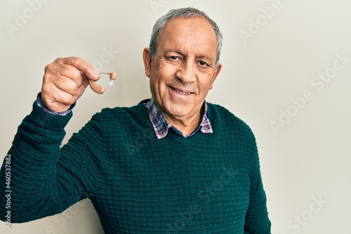 Handsome senior man with grey hair holding medical hearing aid looking positive and happy standing and smiling with a confident smile showing teeth