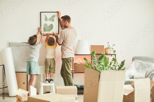 Full length portrait of happy father with two sons hanging pictures on wall while moving in to new home, copy space