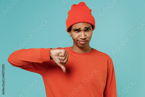 Young sad unhappy displeased african american man 20s in orange shirt hat showing thumb down dislike gesture isolated on plain pastel light blue background studio portrait. People lifestyle concept.