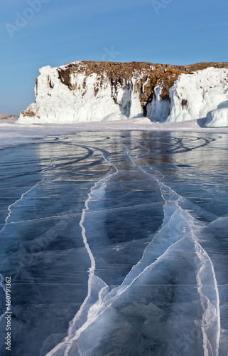 Beautiful winter landscape with blue transparent ice with cracks on a sunny frosty day. In the distance, icy white cliffs of Olkhon Island on frozen Baikal Lake. Unusual natural winter background