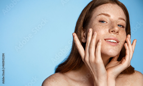 Portrait of redhead female model using skincare product, applying facial cleanser, looking at bathroom mirror, blue background