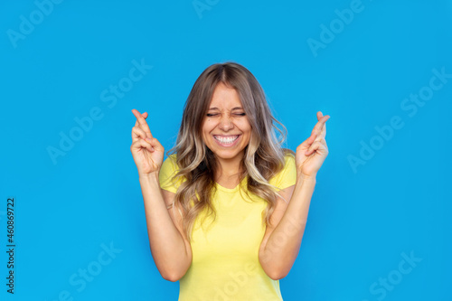 A young pretty smiling blonde woman in a yellow t-shirt with her eyes closed crosses her fingers for good luck waiting for the results of the lottery or exams isolated on a color blue background