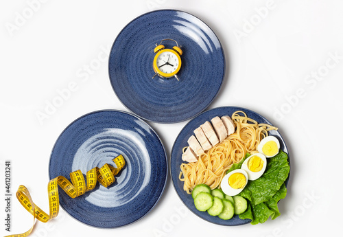 Plates with food, alarm clock and measuring tape, intermittent fasting concept.