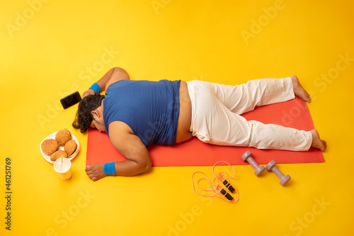 A fat man sleeping on yoga mat amidst dumbles,skipping rope,Burger,Drink and mobile phone.