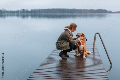 Girl in green jacket sitting on pier with brown Nova Scotia Duck Tolling Retriever and stroking pet. Travelling with dog. Selective focus. Friendship between animals and humans. Foggy background.