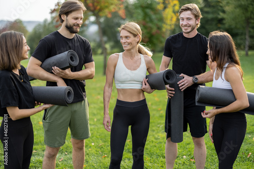 European people with fitness mats standing and talking in green meadow. Friends practicing yoga and doing sport exercises. Concept of healthy lifestyle. Athletic men and women wearing sportswear
