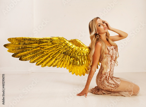 A beautiful young model with big golden angel wings is sitting in the studio. white background