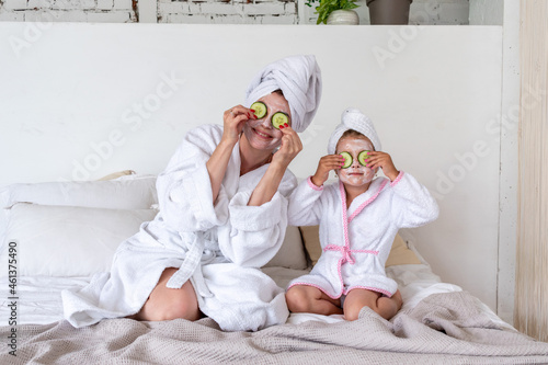 Mom and daughter sitting the bed with cosmetic mask and cucumbers on their faces. Clothes are white terry robes.