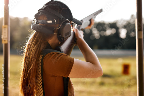 Young caucasian woman on tactical gun training classes. Woman with weapon, wearing cap, protective headphones and eyeglasses. Outdoor Shooting Range. At Sunny evening at summer. Rear view