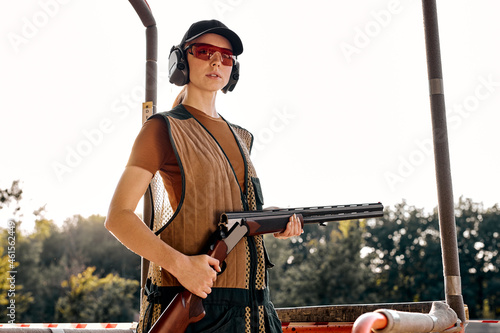 Good-looking caucasian lady with rifle weapon in outdoor academy shooting range, field in the background. Young beautiful woman in cap, protective headset and spectacles posing, ready to train