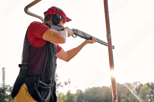 Side View on Young Caucasian Man wear Ear Plug Confidently Aiming Shotgun At Target in Outdoor Shooting Range, Alone. Man Practicing Fire Weapon Shooting. Hobby, Skill, Shooting Concept.