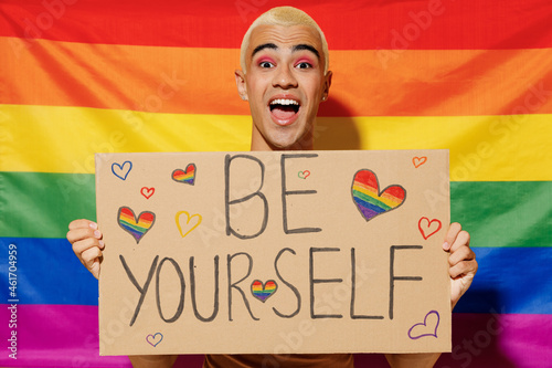 Young queer bisexual surprised blond latin gay man with make up in beige tank shirt hold card sign with be yourself title text on rainbow flag background studio portrait People lgbt lifestyle concept.