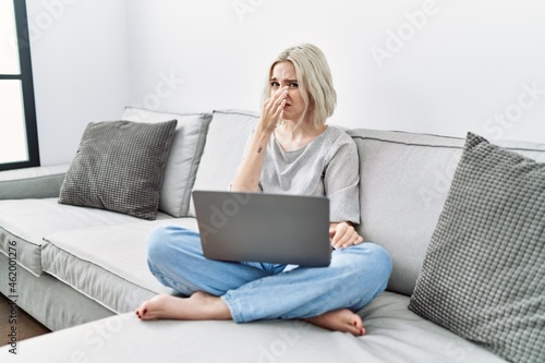 Young caucasian woman using laptop at home sitting on the sofa smelling something stinky and disgusting, intolerable smell, holding breath with fingers on nose. bad smell