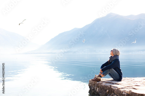 Young woman relaxing at winter sea beach. Traveler resting by blue mountain