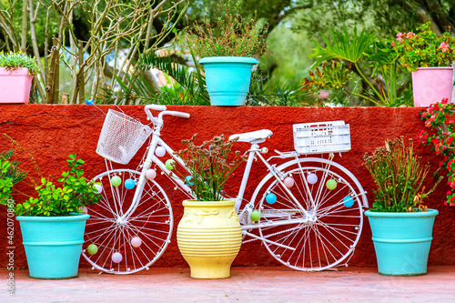 Old white bicycle near colorful flower pots against red wall in Greece.