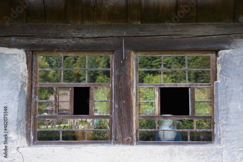 Doppelfenster eines traditionellen Berghofs im Schwarzwald. Die Fenster sind handgefertigt aus Holz, die kleinen rechteckigen Glasscheiben reflektieren den Wald. Die Mauern sind weiß verputzt. 