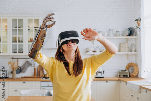 Positive young woman in casual clothes with modern bionic arm and virtual reality headset dances near sofa in stylish kitchen