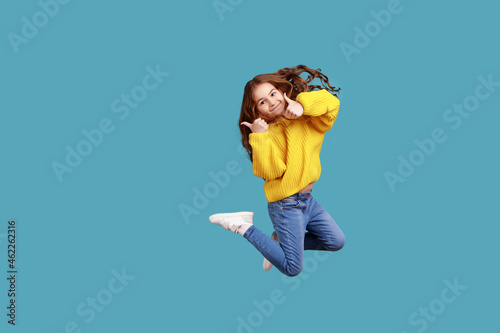 Full length portrait of happy charming little girl jumping up high and showing thumb up to camera, wearing yellow casual style sweater. Indoor studio shot isolated on blue background.