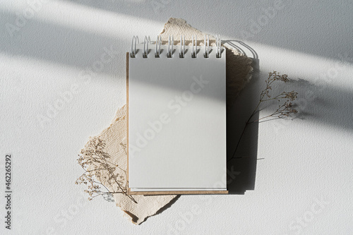 Notepad mockup for showcasing artwork and design. A notebook with an open blank page lies on a white table surrounded by dry plants. Minimal mock up of sketchbook or drawing paper.