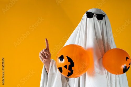 Person in Halloween costume of ghost with sunglasses and Jack-o'-lantern balloons points away