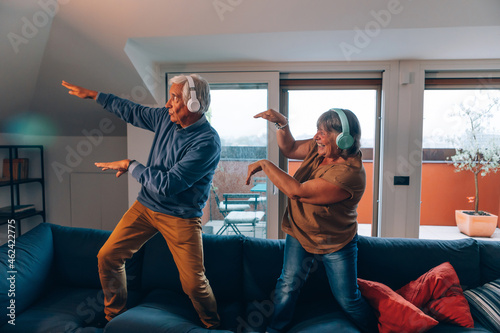 happy active senior couple plays and dances on sofa at home with headphones listening to music