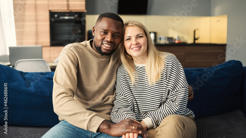 Portrait of beautiful diverse couple of Black man and Caucasian woman looking at camera and smiling sitting on sofa at home. Loving biracial couple posing in embrace
