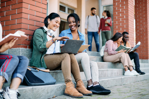 Happy multi-ethnic female students have fun while using laptop in front of university building.