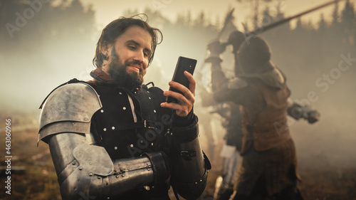 Portrait of Handsome Medieval Knight Using Smartphone on Battlefield, Smiling. Funny Concept: Armored Warrior Having Fun, Ordering Online, Betting, Investing, doing E-commerce. War is Raging