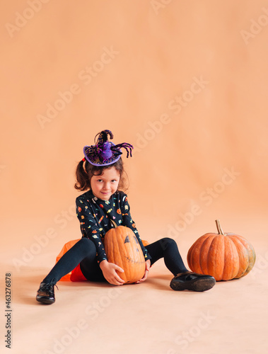 Funny little girl in a witch costume and a hat with a spider for Halloween with a pumpkin Jack on a plain background