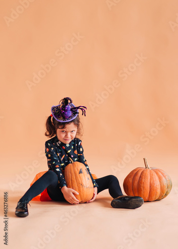 Funny little girl in a witch costume and a hat with a spider for Halloween with a pumpkin Jack on a plain background